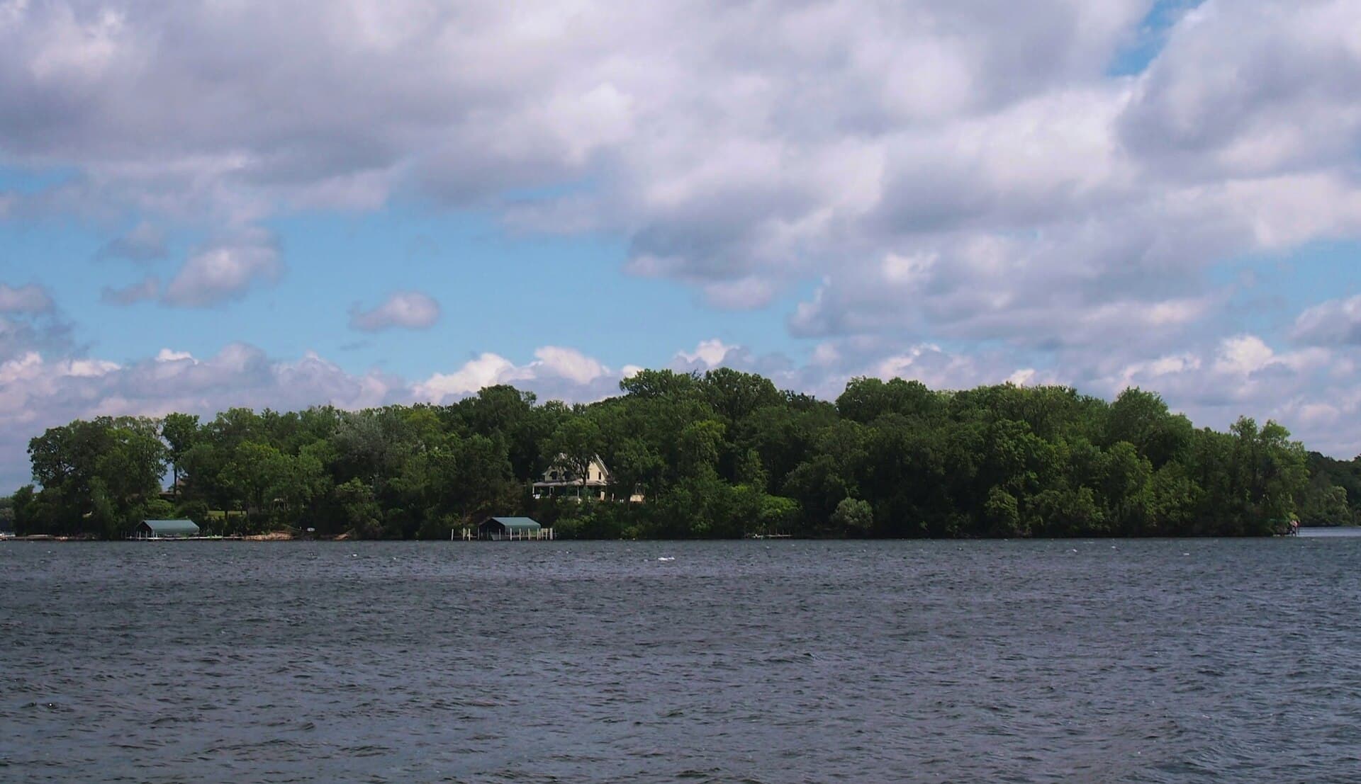 Crane Island seen across Lake Minnetonka from the boat launch at Lake Minnetonka Regional Park in Minnetrista, wooded shoreline and a lakeside home under a bright, cloud-dotted summer sky