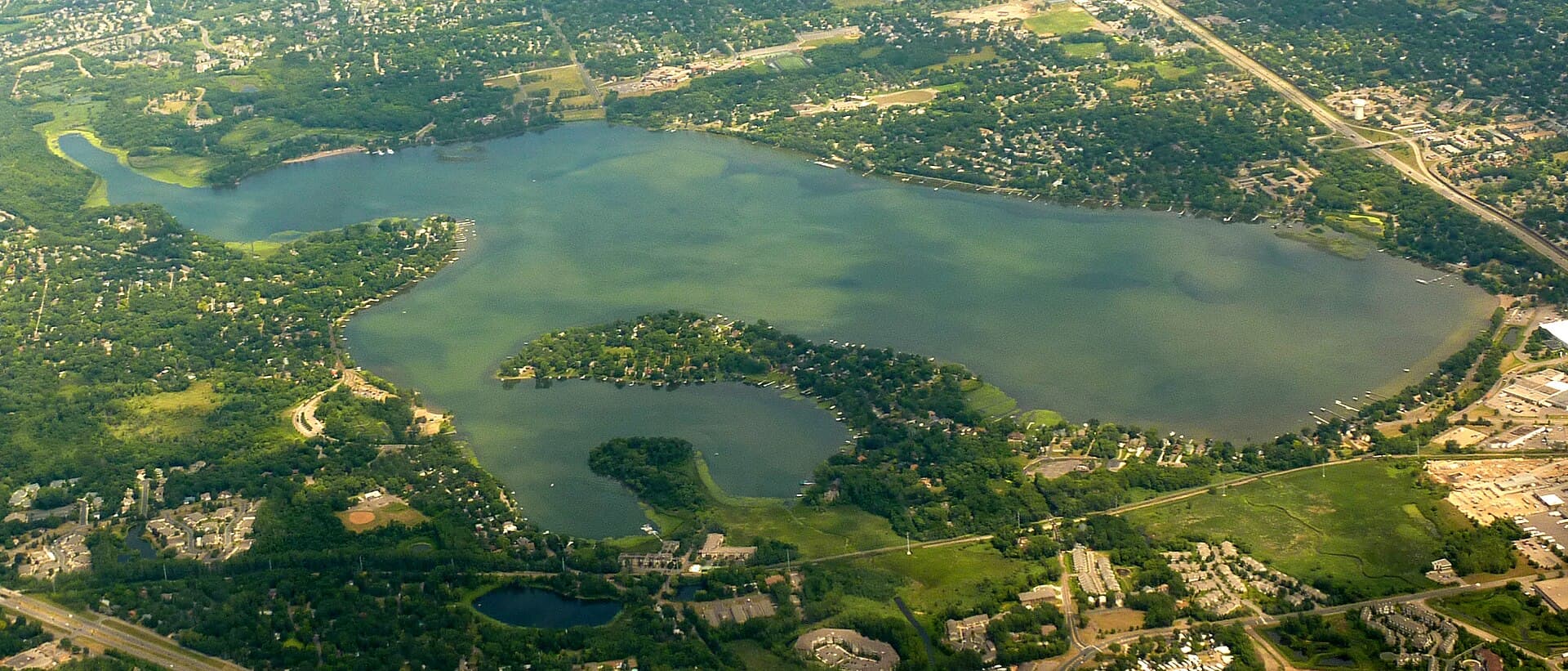Aerial view of Medicine Lake in Plymouth, the green wooded peninsula jutting into the lake and ringed by leafy residential neighborhoods on a sunny day